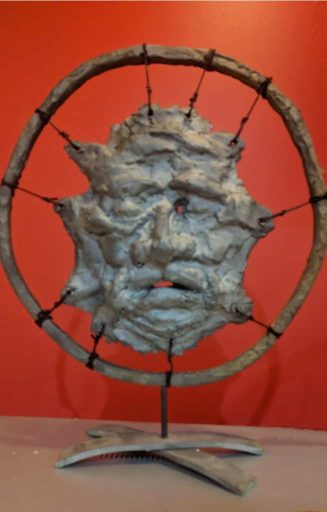 Intricate carved face sculpture supported by a circular wooden frame against a vibrant orange background, showcasing detailed craftsmanship and artistic expression.
